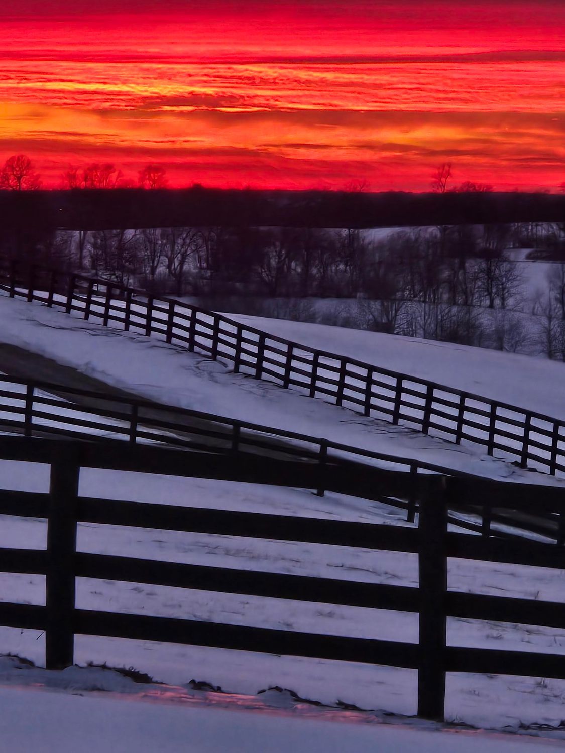 A sunset over a snowy field with a fence in the foreground