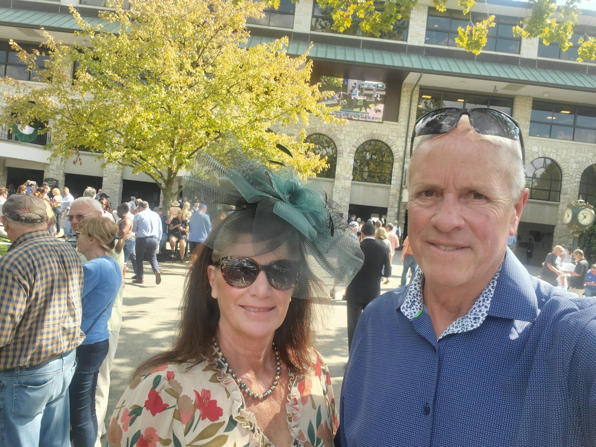 A man and a woman are posing for a picture at a race track . the woman is wearing a hat and sunglasses.