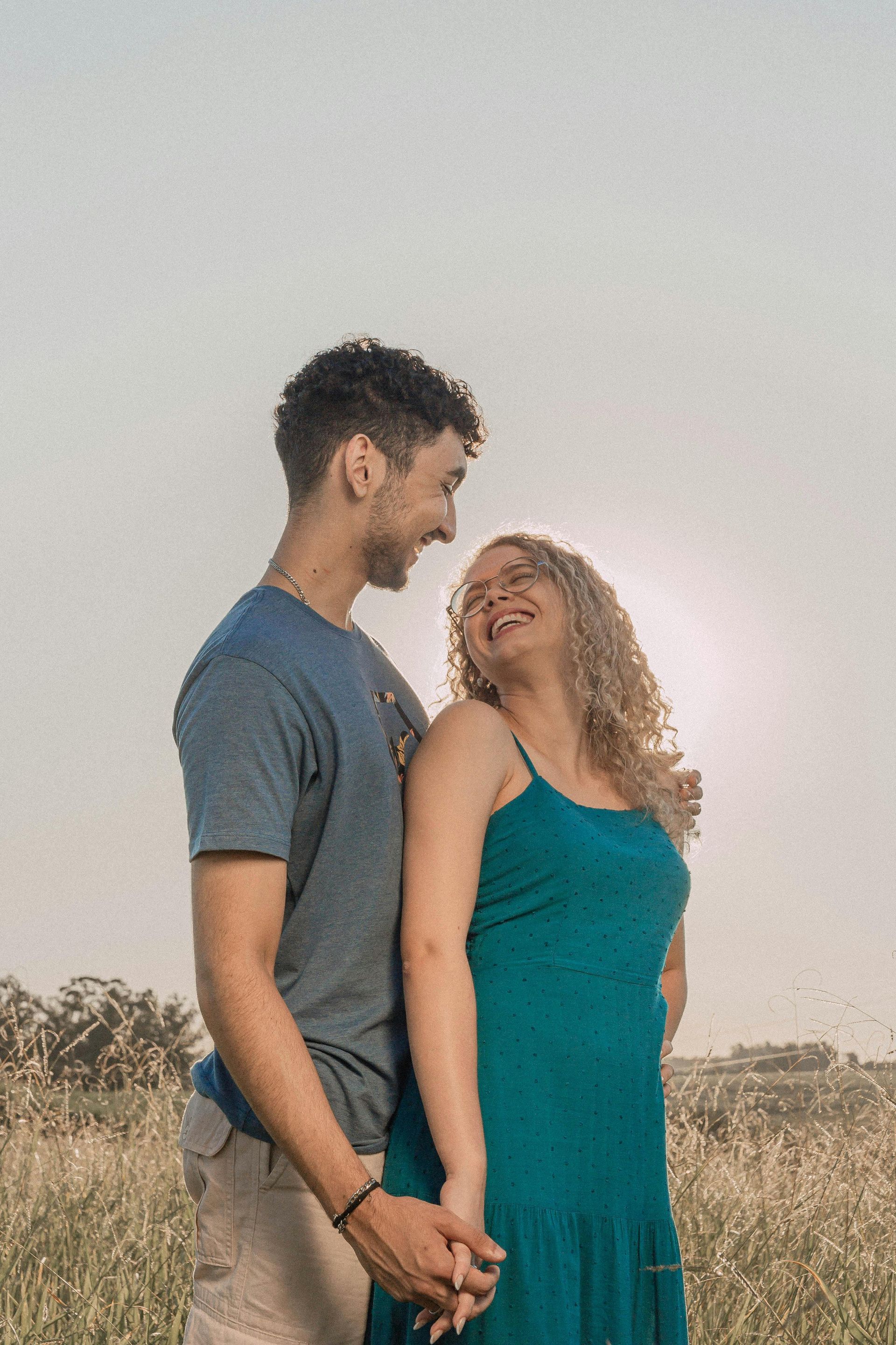 Couple holding hands, laughing, looking at each other in a field. The sun is behind them.