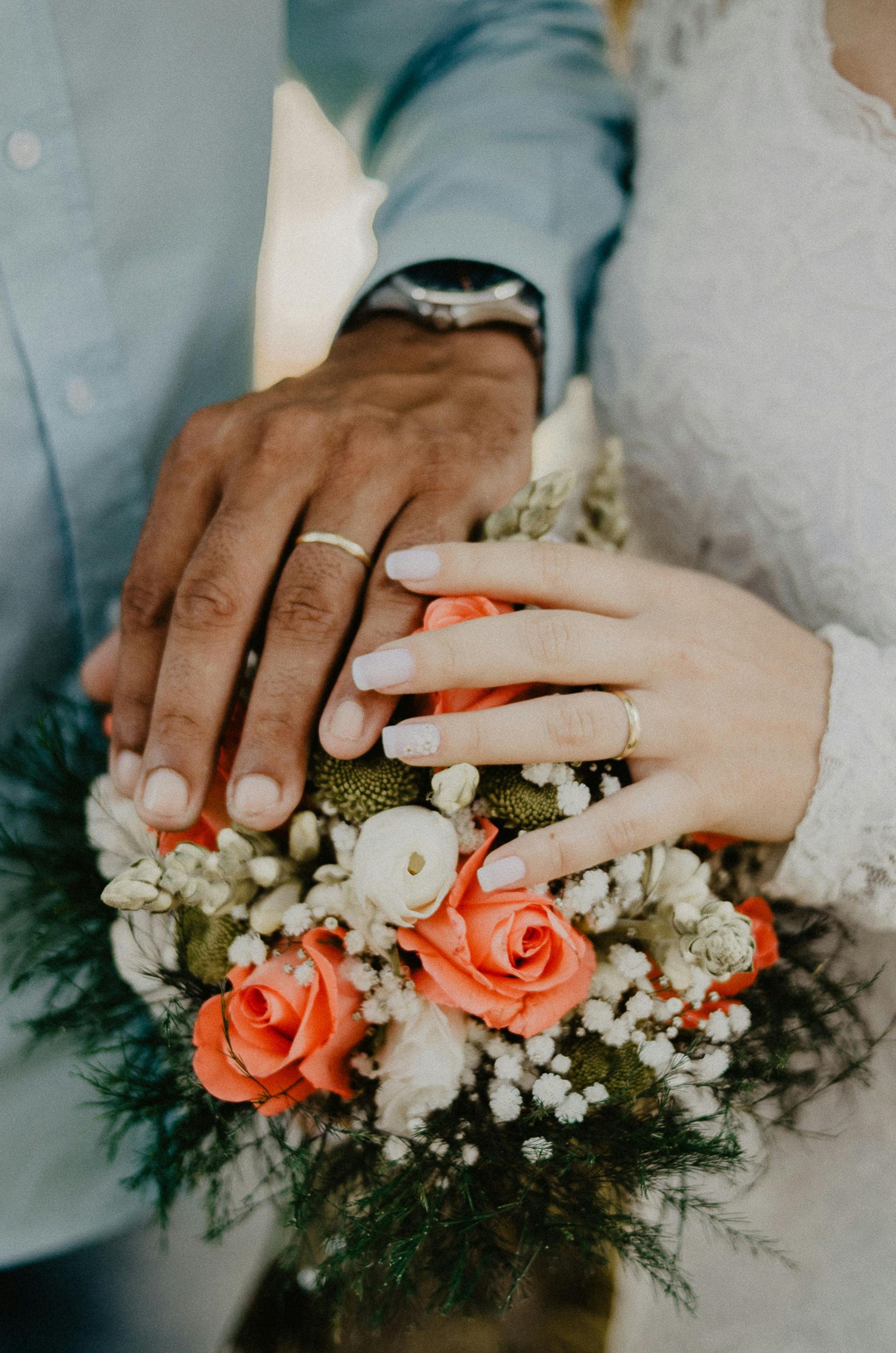 A close up of a bride and groom 's hands with wedding rings holding a bouquet of flowers.
