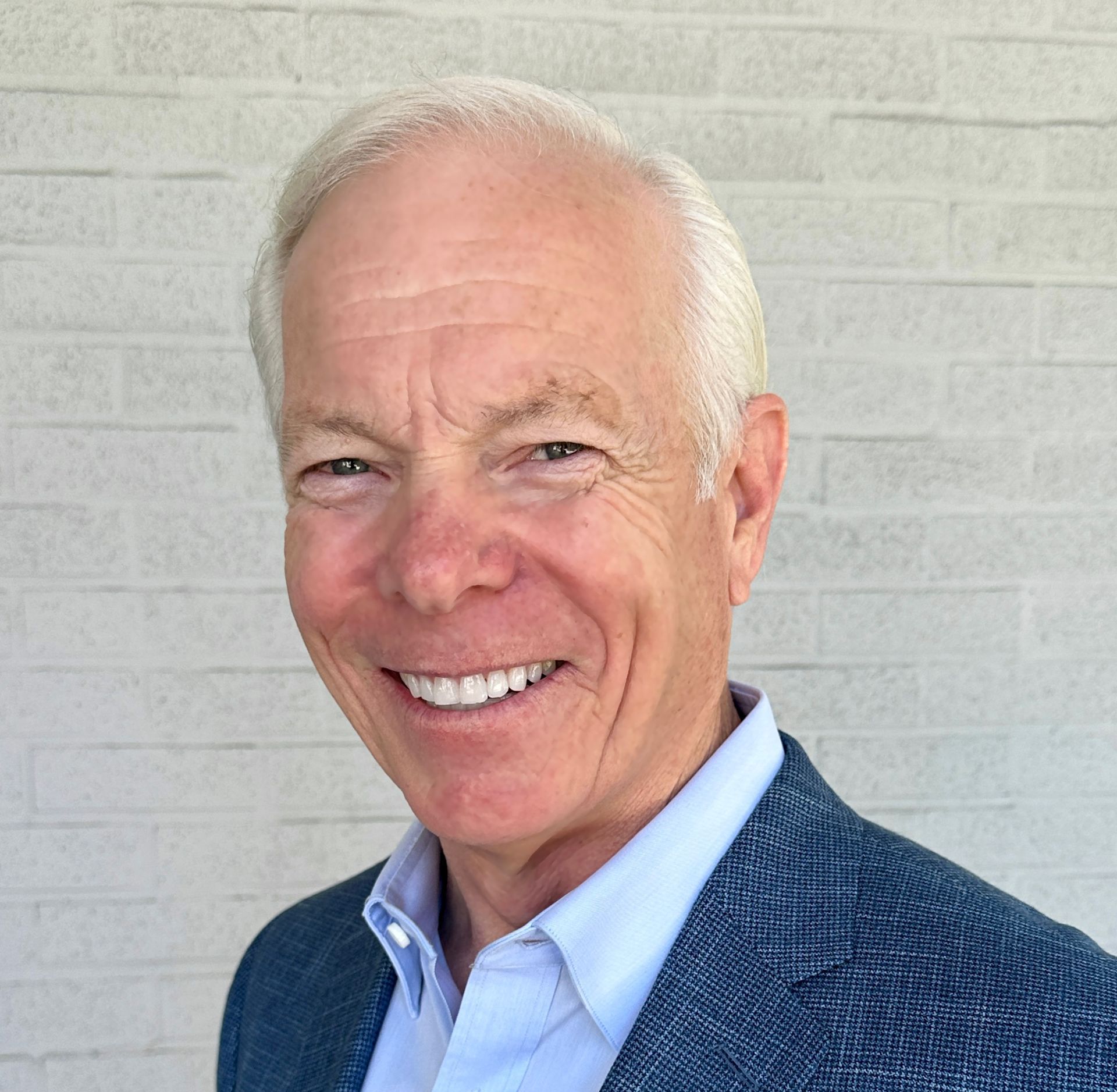 A man in a suit and white shirt is smiling in front of a white brick wall.