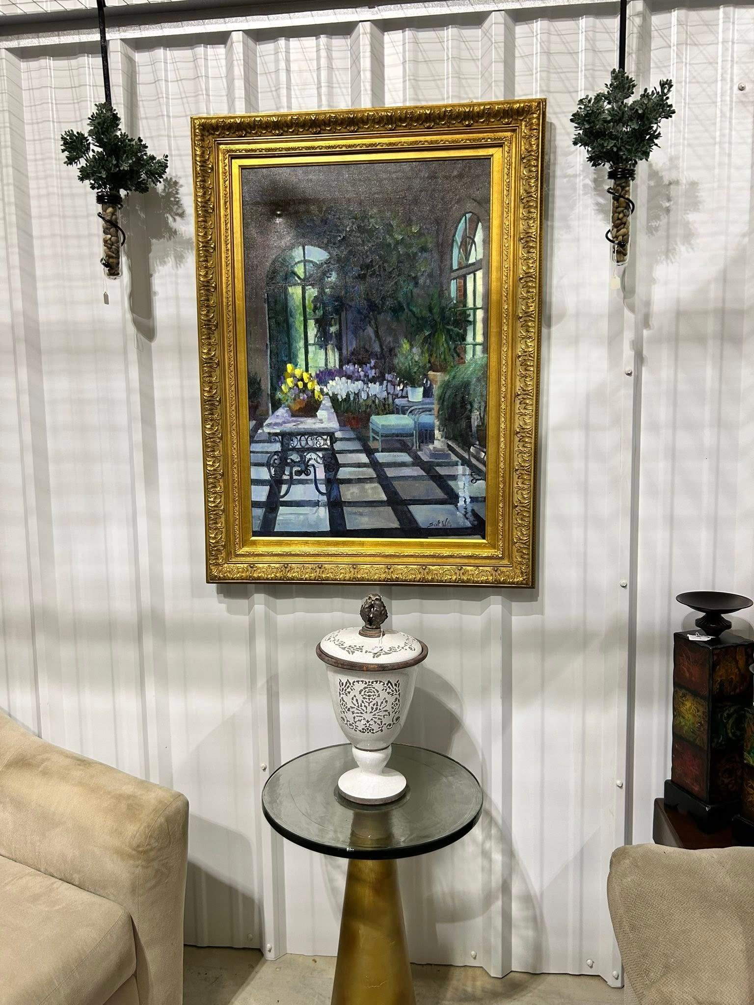 A framed painting of a garden, over a table with a decorative vase, between two chairs, with greenery on either side.