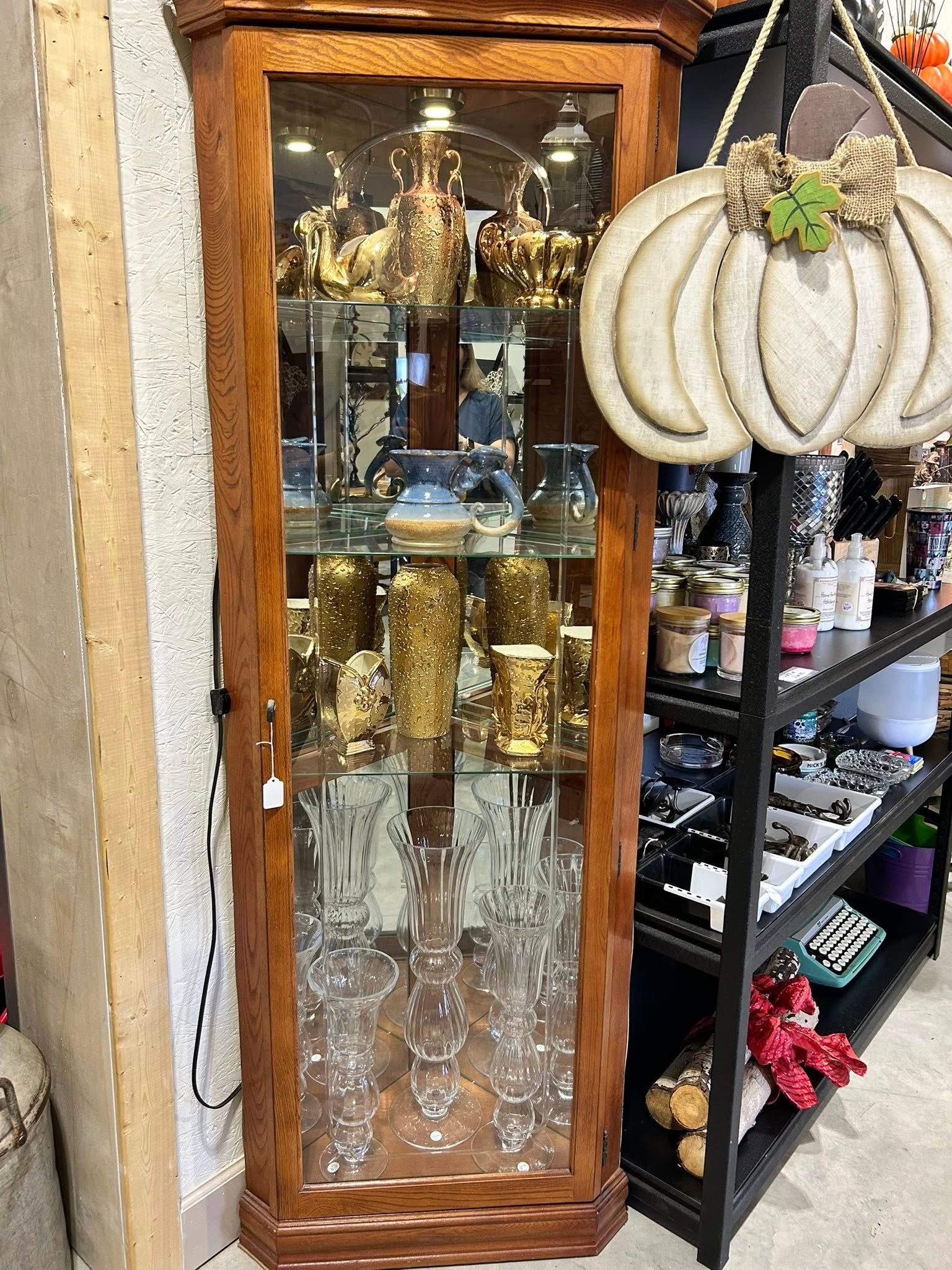Wooden curio cabinet filled with gold vases and etched glassware. A white pumpkin decoration hangs nearby.