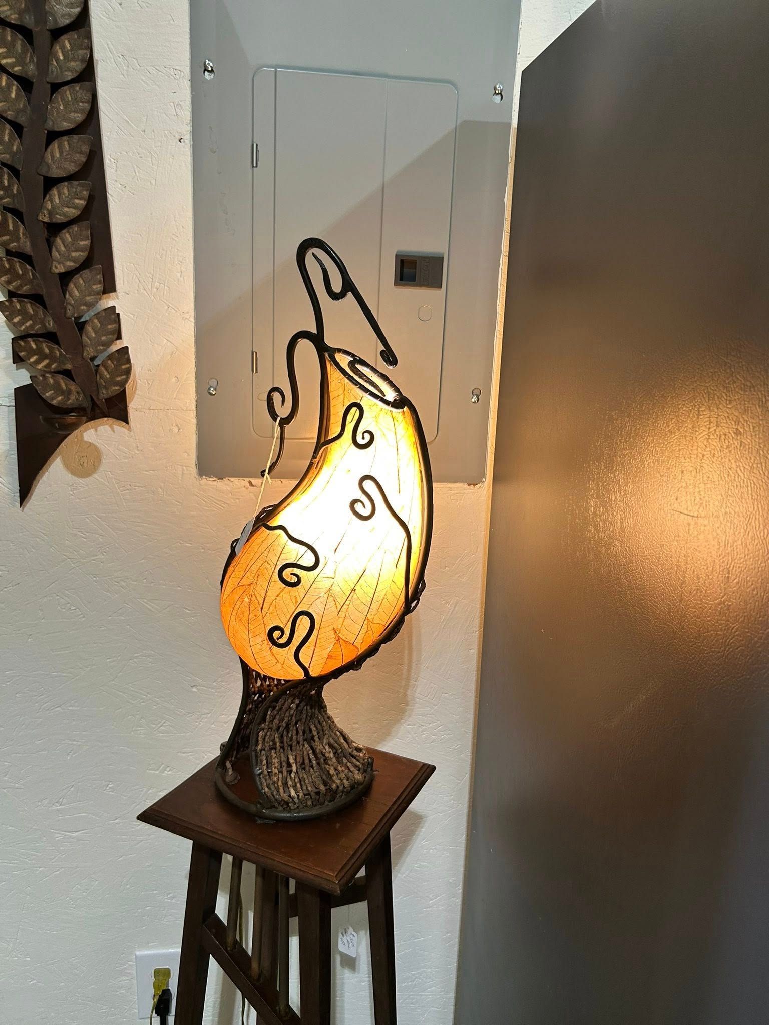 Decorative lamp on wooden stand, illuminating a wall with a leaf art piece and electrical panel.