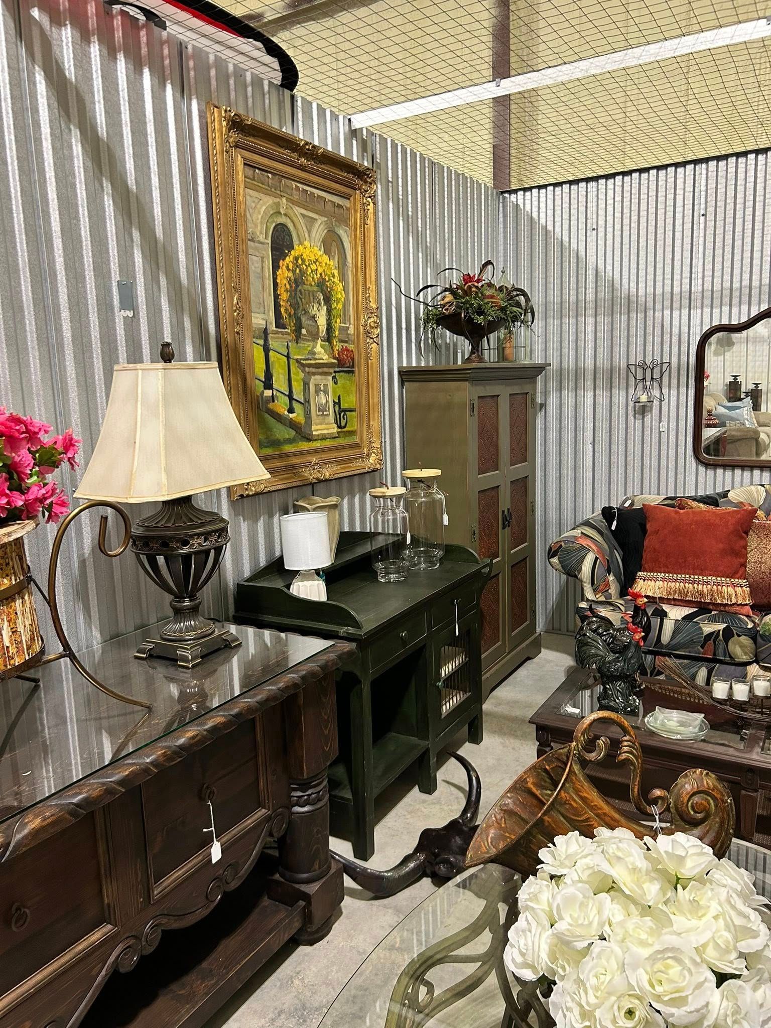 Antique furniture display in a corrugated metal-walled room, featuring a lamp, painting, and cabinet.