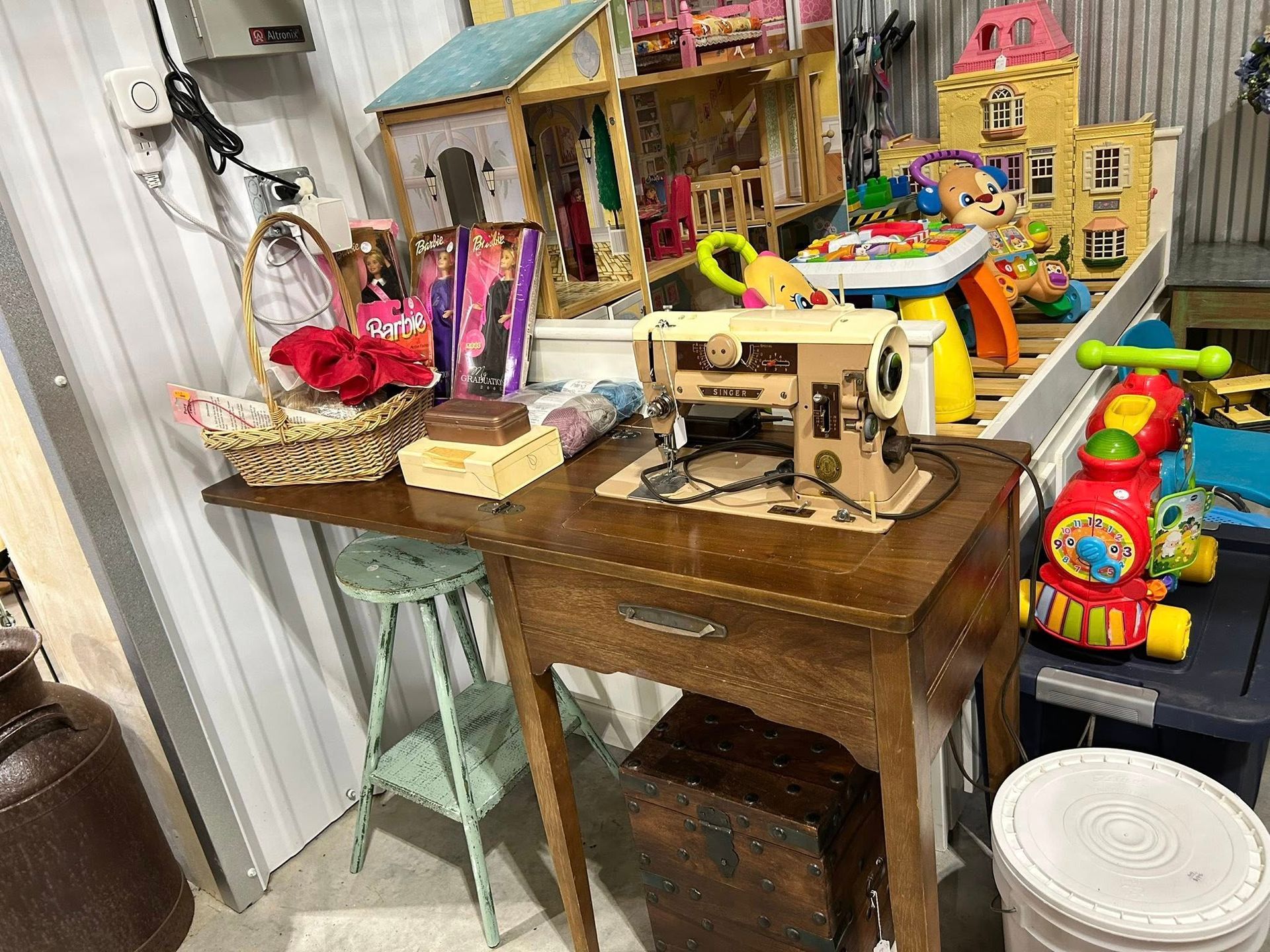 A cluttered storage room with a sewing machine on a wooden table. Toys and other items are piled around.