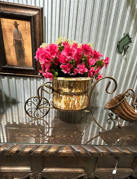 Metal bicycle planter with pink flowers on a wooden table, framed artwork in the background.