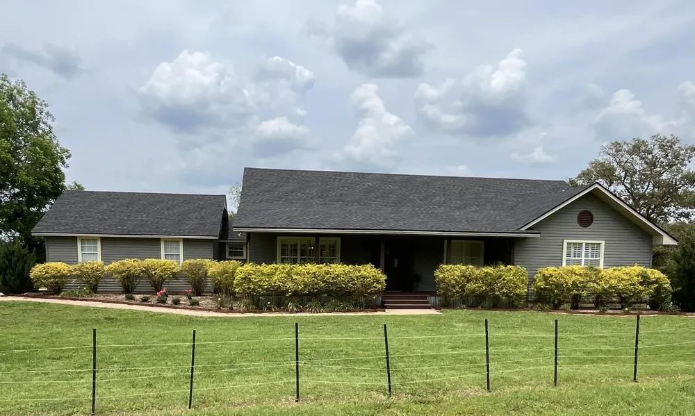 Gray house with dark gray roof and green bushes in front, on a grassy field under cloudy sky.