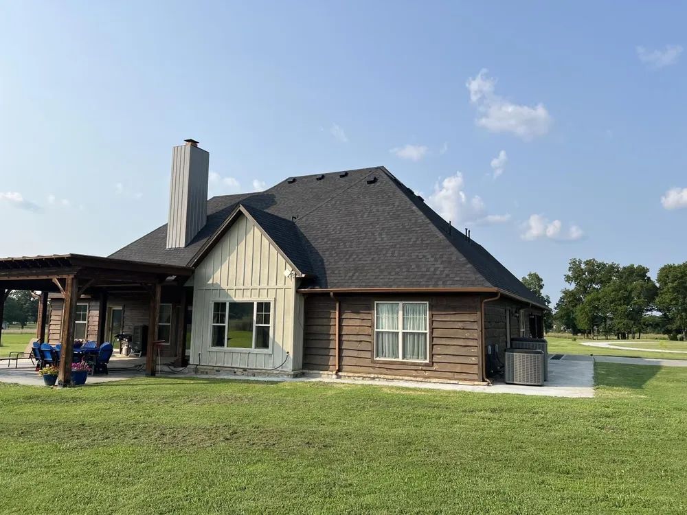 Brown house with dark roof, chimney, windows, and attached pergola on a grassy lawn under a blue sky.