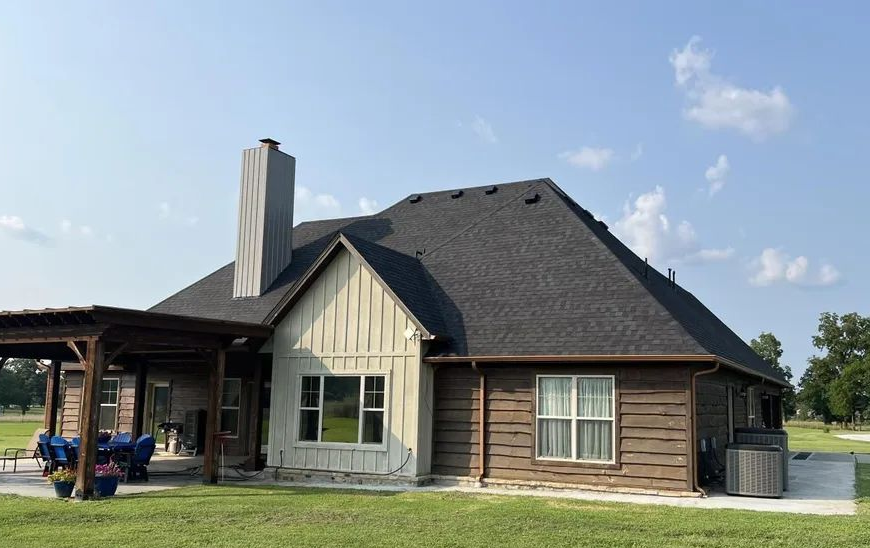 A view of the back of a house featuring brown wood siding, a cream-colored board and batten section, and a patio area.