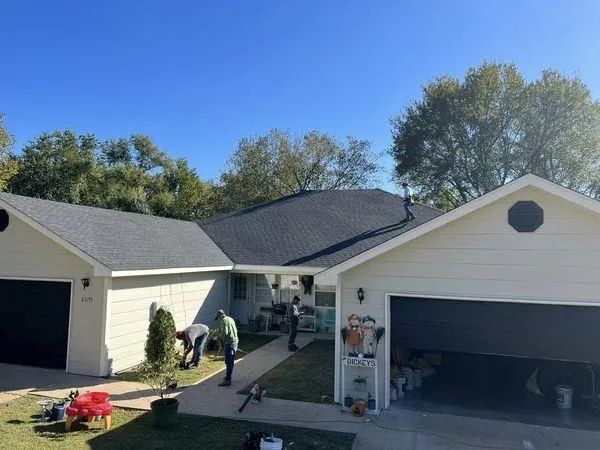 Workers landscaping a house with a new black roof and two black garage doors under a clear blue sky.