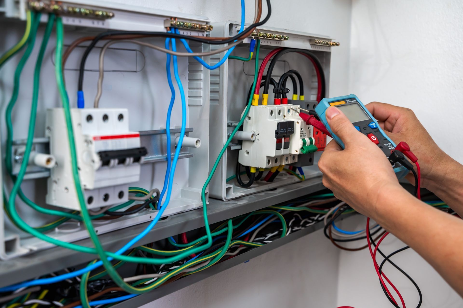 Electrician using a multimeter to inspect wiring in an electrical panel, with various colored wires and circuit breakers.