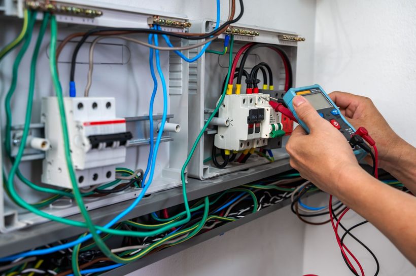 Electrician using a multimeter to inspect wiring in an electrical panel, with various colored wires and circuit breakers.
