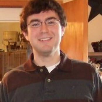 Man with glasses smiles, wearing a brown and gray striped collared shirt.