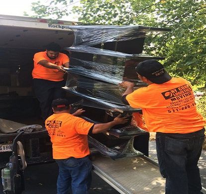 Three movers in orange shirts loading wrapped furniture into a truck.