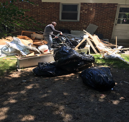 A person sorting debris, including black trash bags and construction waste, in front of a brick building.