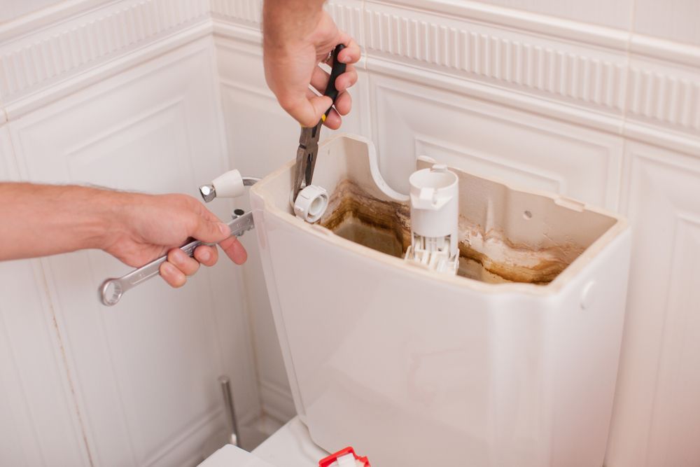 Person repairs toilet tank with wrench and pliers. Inside tank shows stains and components.