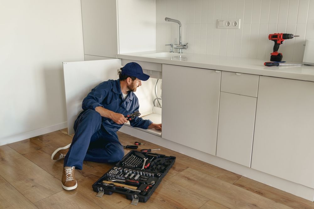 Plumber in blue overalls works under a kitchen sink. A toolbox and drill rest on the floor nearby.