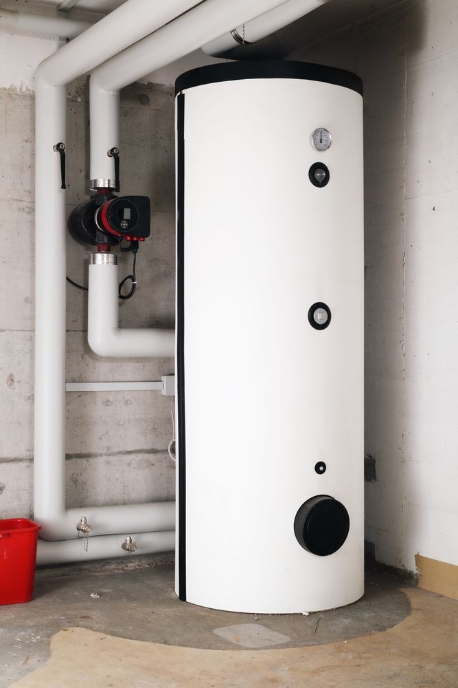 White cylindrical water heater in a basement with pipes and a red bucket.
