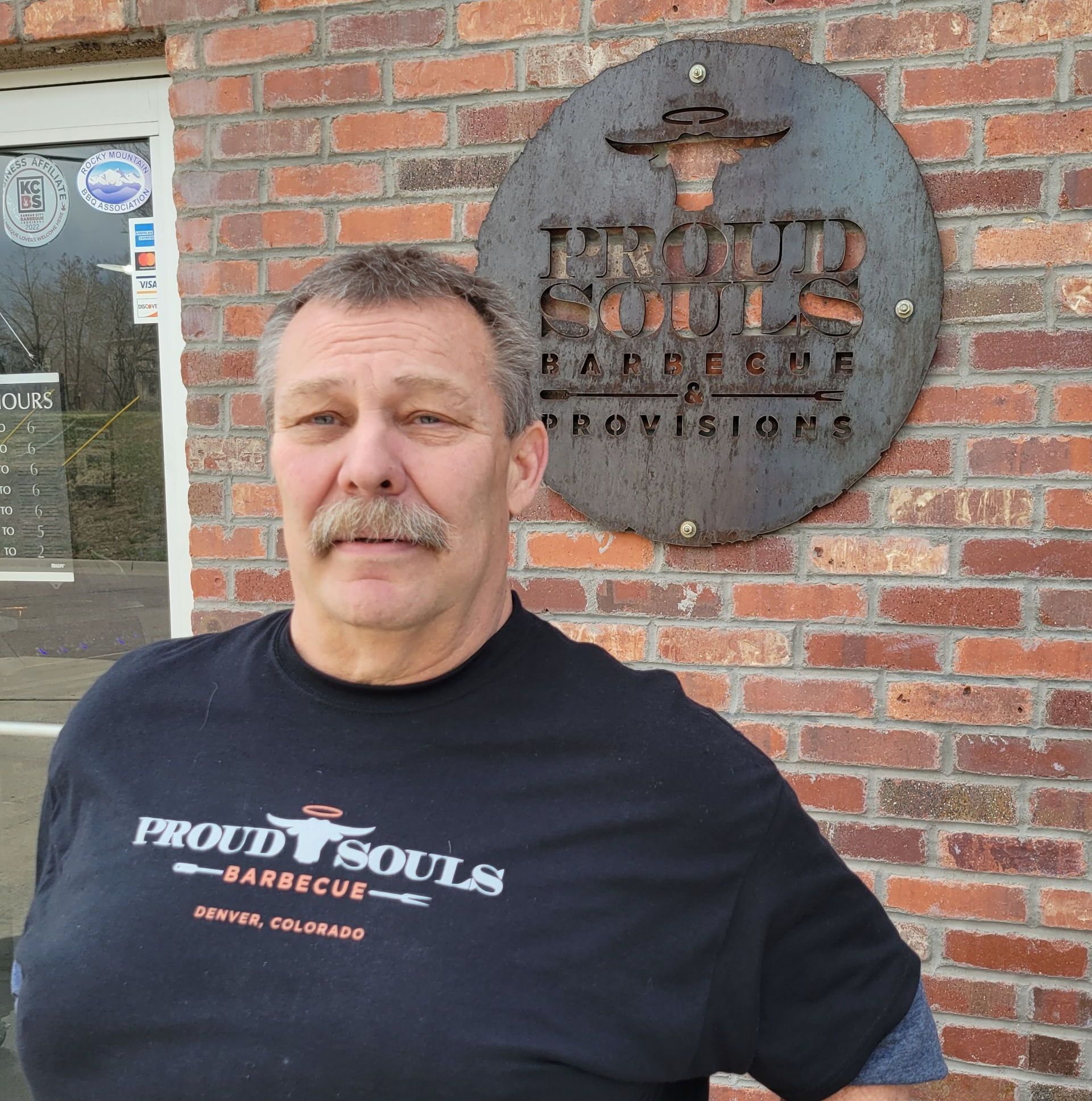 A man wearing a proud souls barbecue shirt stands in front of a brick building