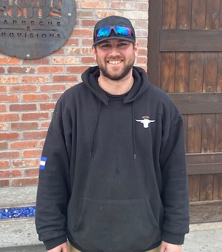 A man wearing a black hoodie and sunglasses stands in front of a brick wall