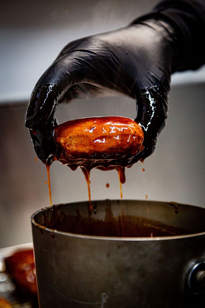 A person in a black glove is holding a piece of meat over a pot of sauce.