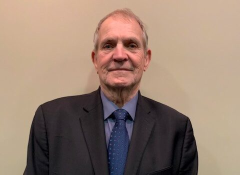 Man in dark suit and blue tie smiles, standing in front of a neutral wall.