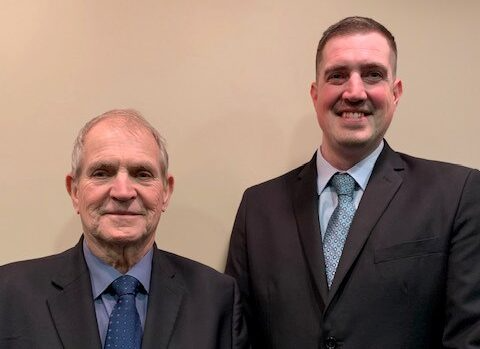 Two men in suits, smiling. Left: older, blue tie. Right: younger, blue tie. Beige wall background.