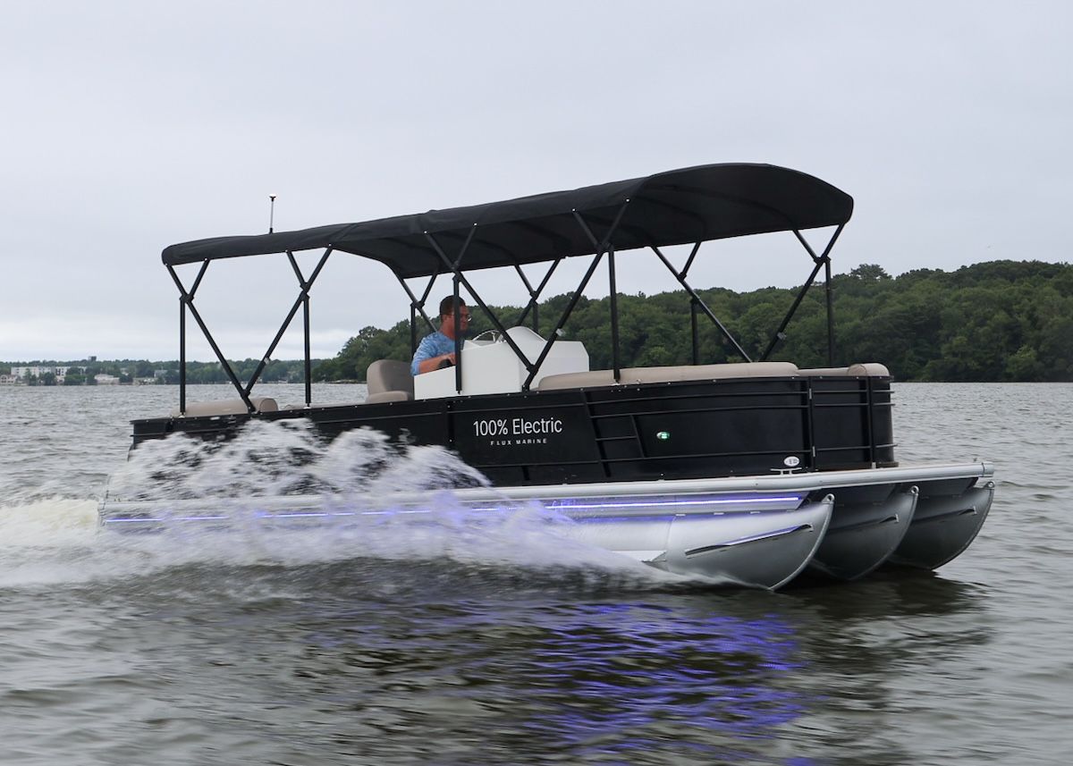 100% electric decoy on side of tritoon that is a black hull tan interior and black bimini up over the seating area. There is a man in the helm driving the boat