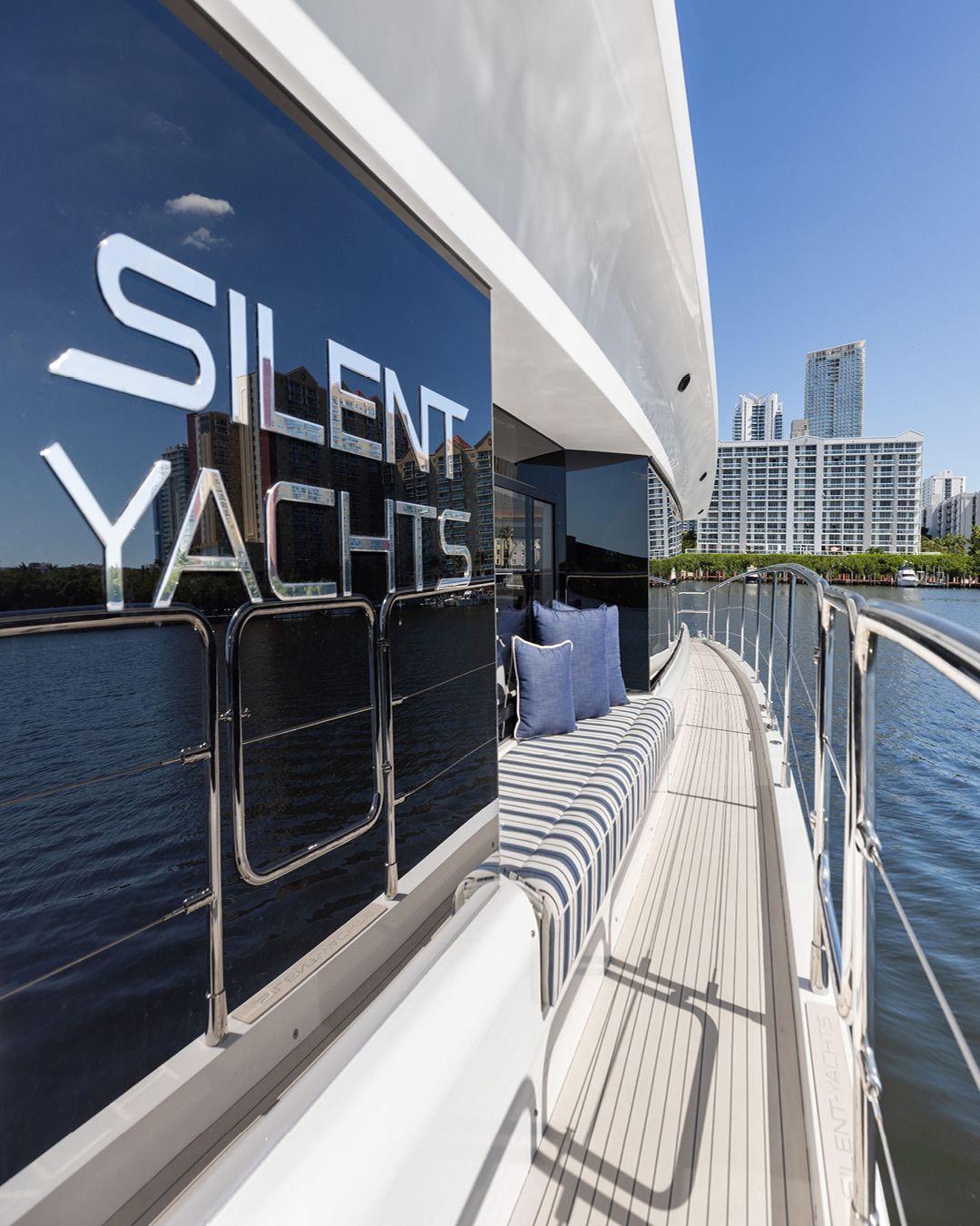 edge of deck on silent yacht boat with striped bench and blue pillows there is a building in the background and water to the right