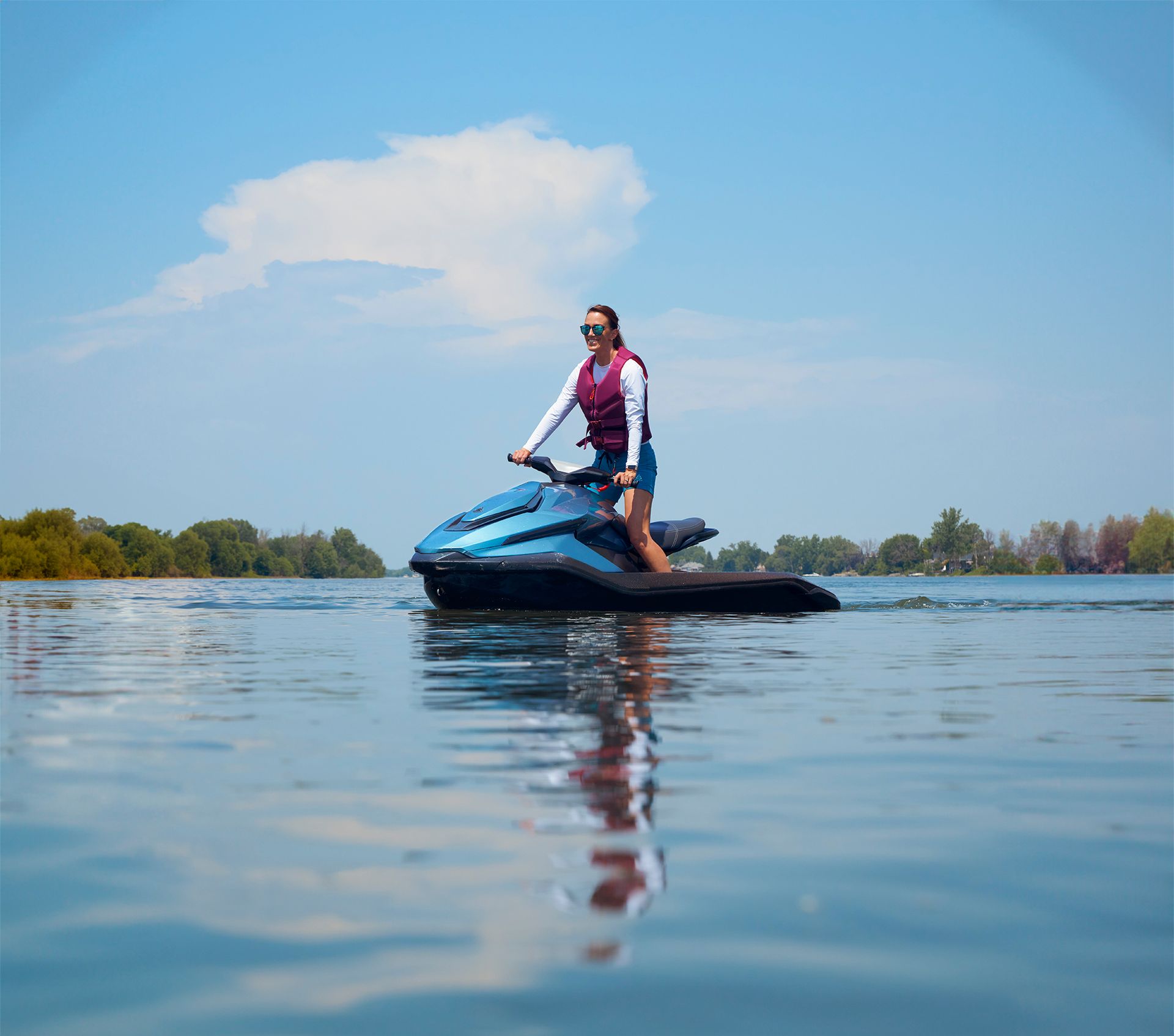 woman in open water standing on platform of blue electric Jetski from Taiga Orca. There are trees and blue sky in the background