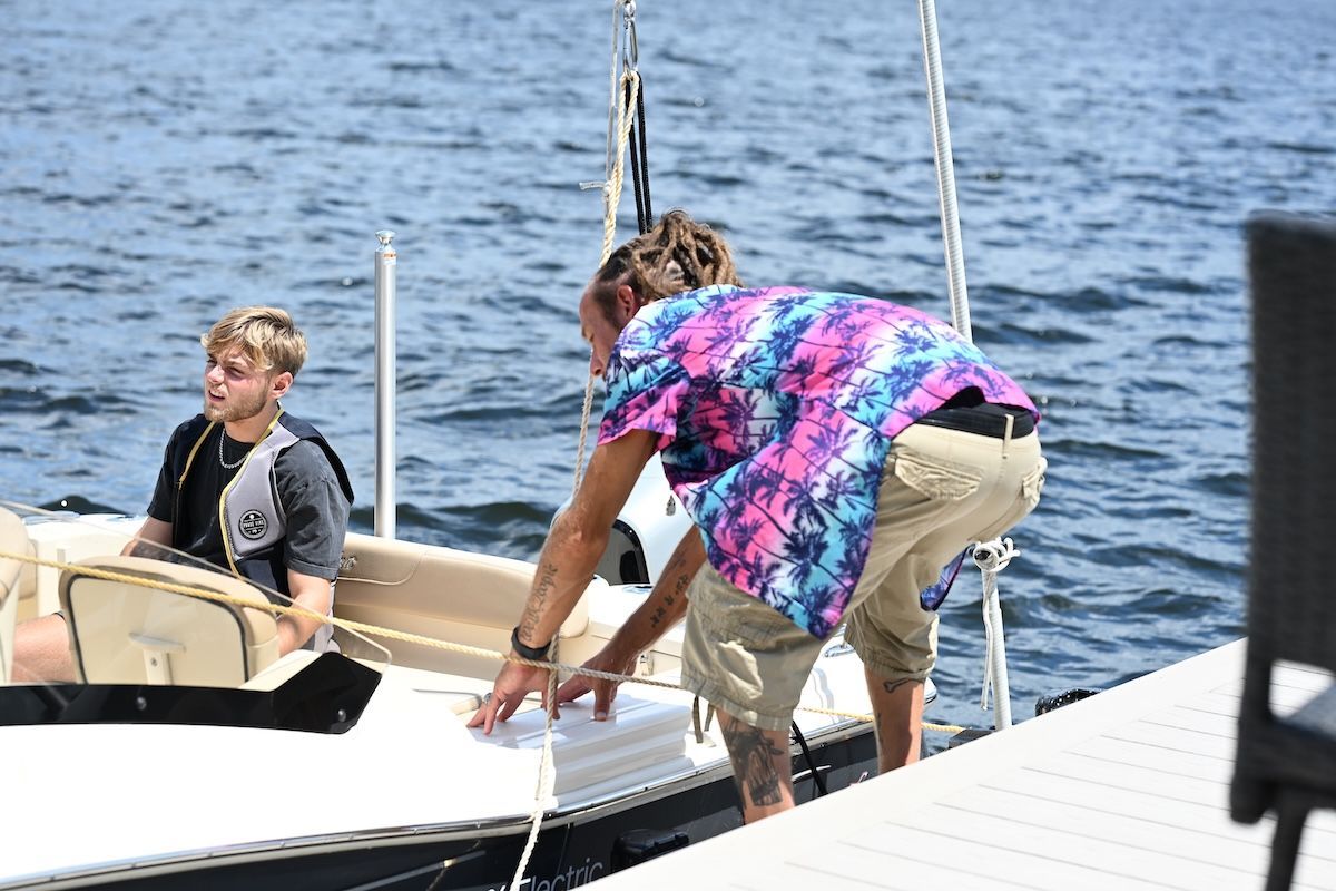 Man pushing boat off dock so it goes out in water wearing bright pink and blue shirt. 