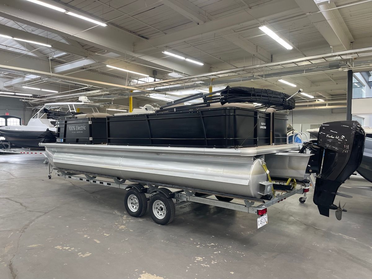 Flux Capacitor tritoon black boat sitting on trailer in warehouse there is a  black outboard motor on the back of it