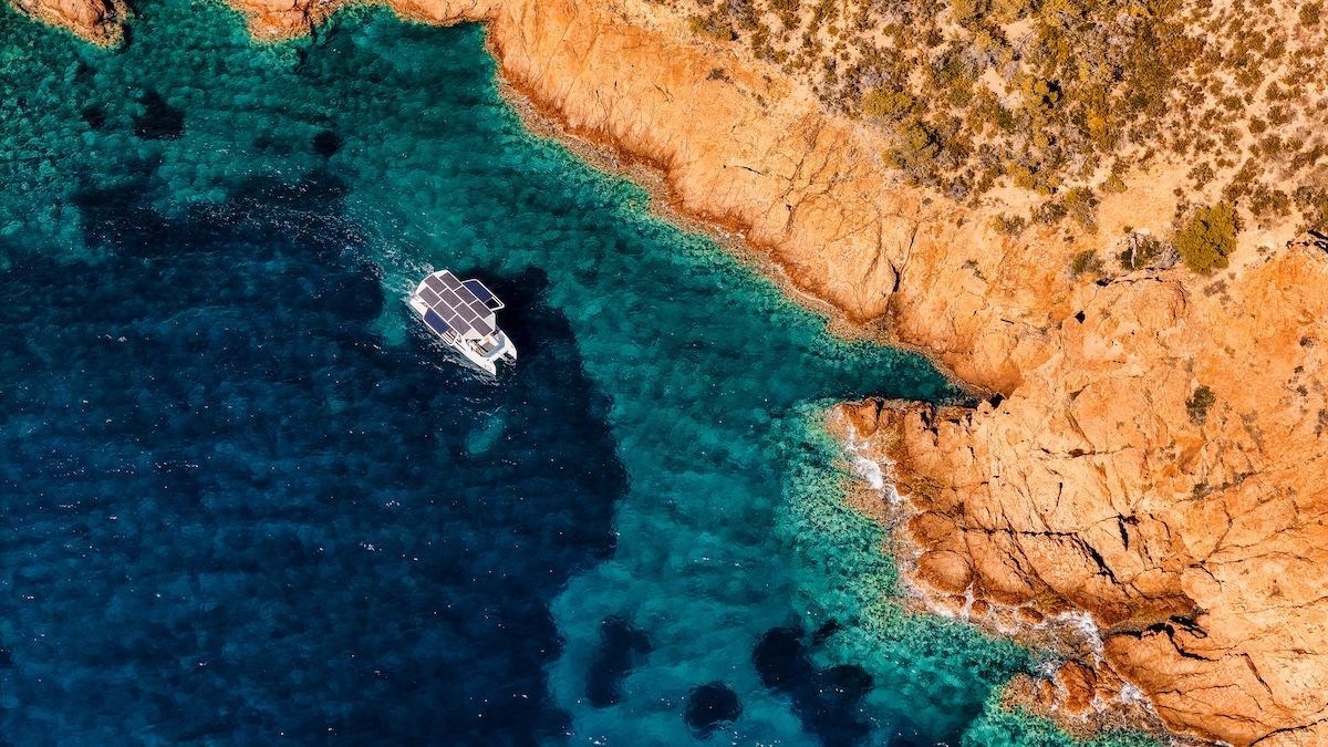 Aerial shot of solar catamaran white with open wings in turquoise blue water and red/brown rock or terrain next to it
