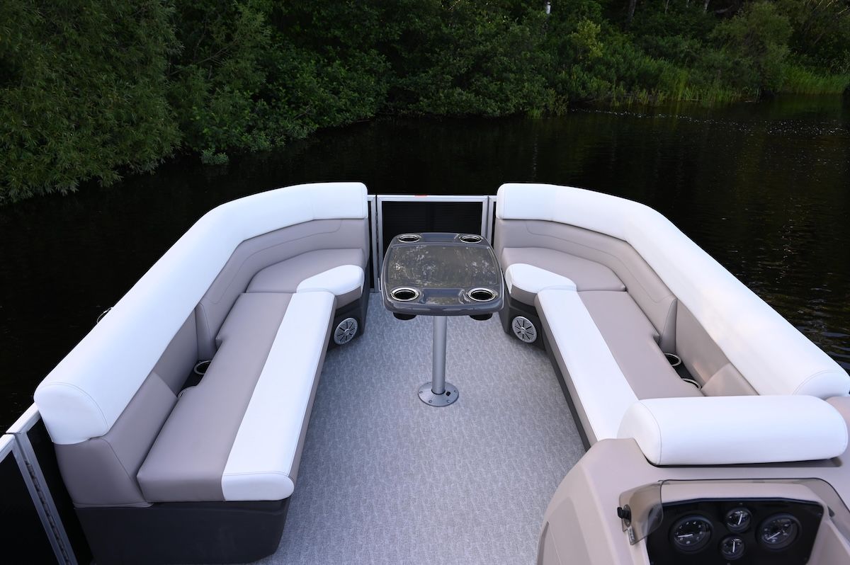 bow of a pontoon, side seating with gate in the front and table set up also trees in the background the cushions on the seats are beige and white