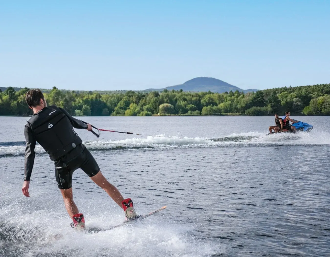 man wakeboarding behind blue electric jetski that is holding two people trees and mountains in background