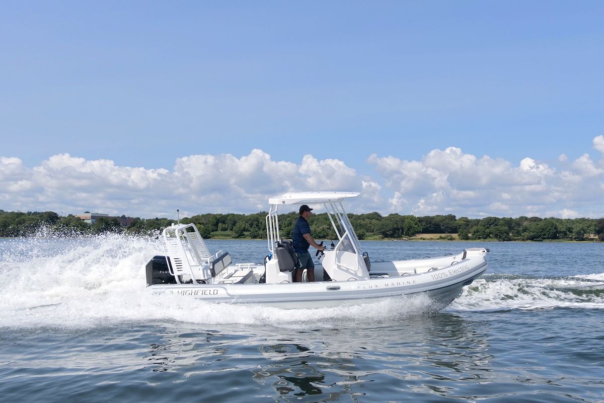 man in white Highfield RIB with black Flux motor on the back. Blue sky and clouds in sky. Trees in background. Man is wearing a baseball hat and a polo shirt