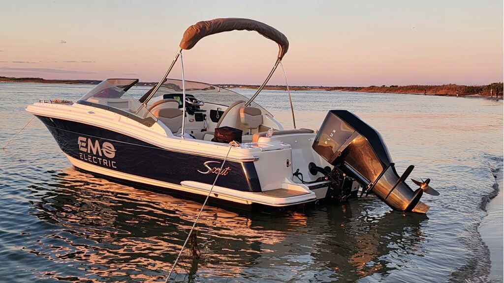 A blue bowrider is sitting in the water near the shore of the  beach at sunset with a black motor on the back of it