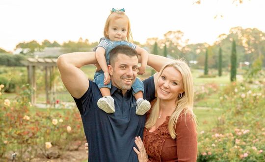 A man and woman are holding a baby on their shoulders in a park.