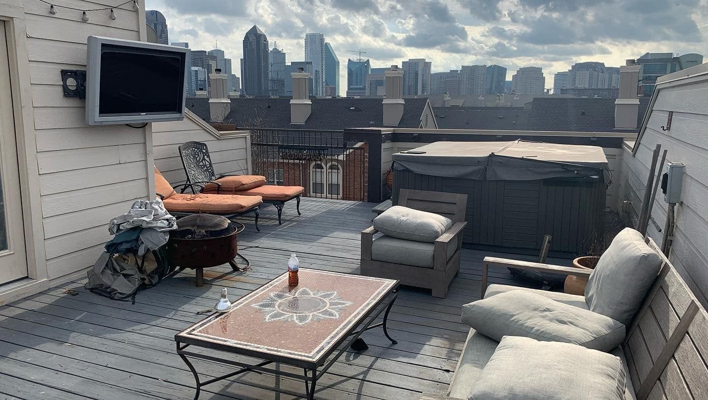 Rooftop patio with seating, fire pit, TV, and hot tub; city skyline in the background under cloudy sky.