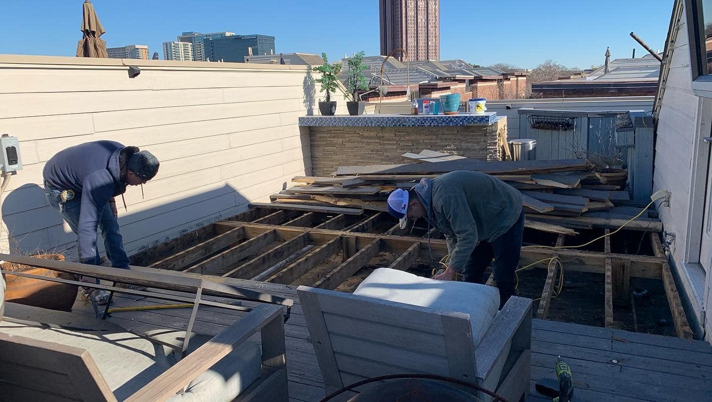 Two people repair rooftop deck with exposed beams; urban setting on a sunny day.