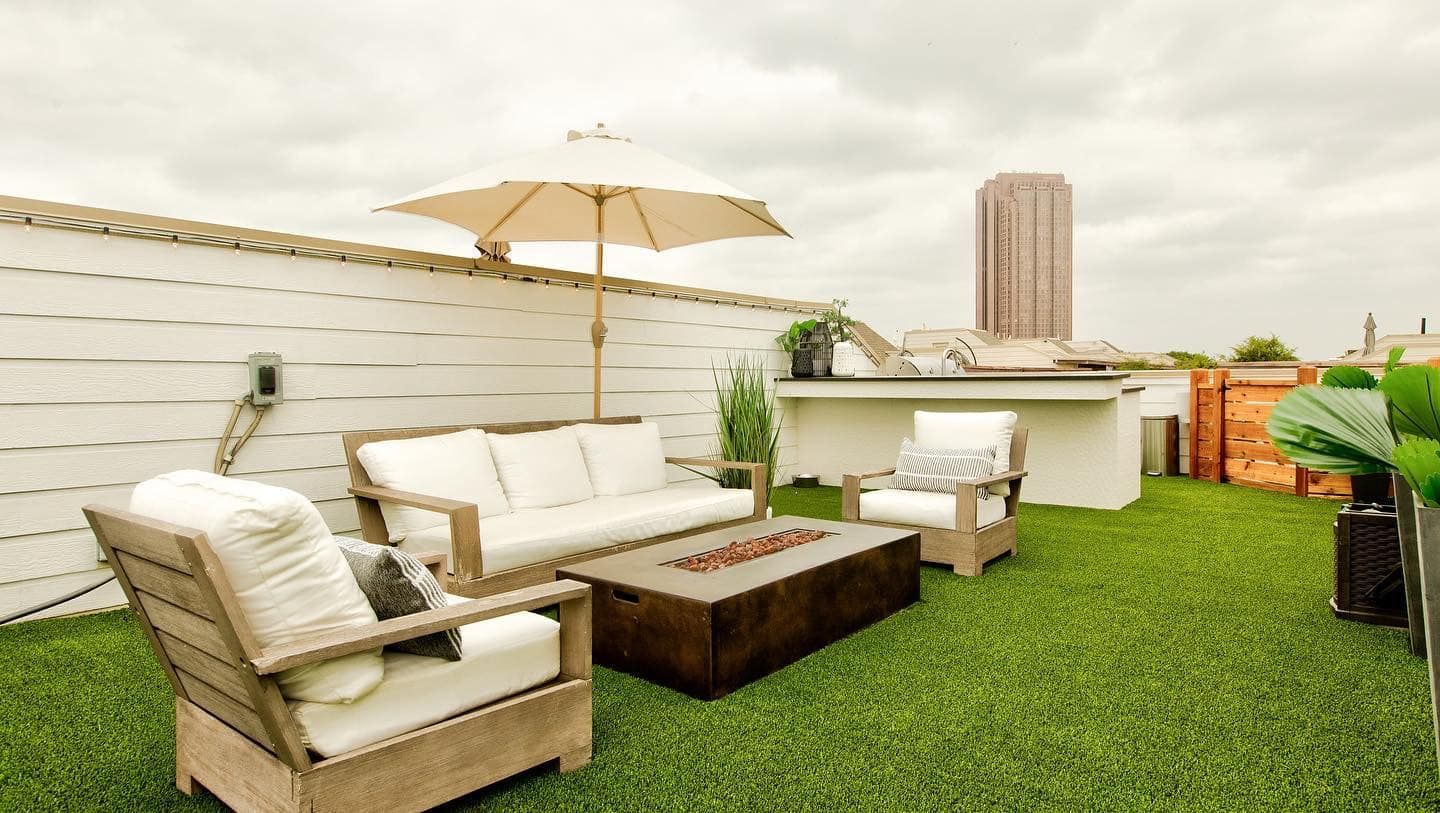Rooftop patio with white furniture, fire pit, umbrella, and a city skyline backdrop on a cloudy day.