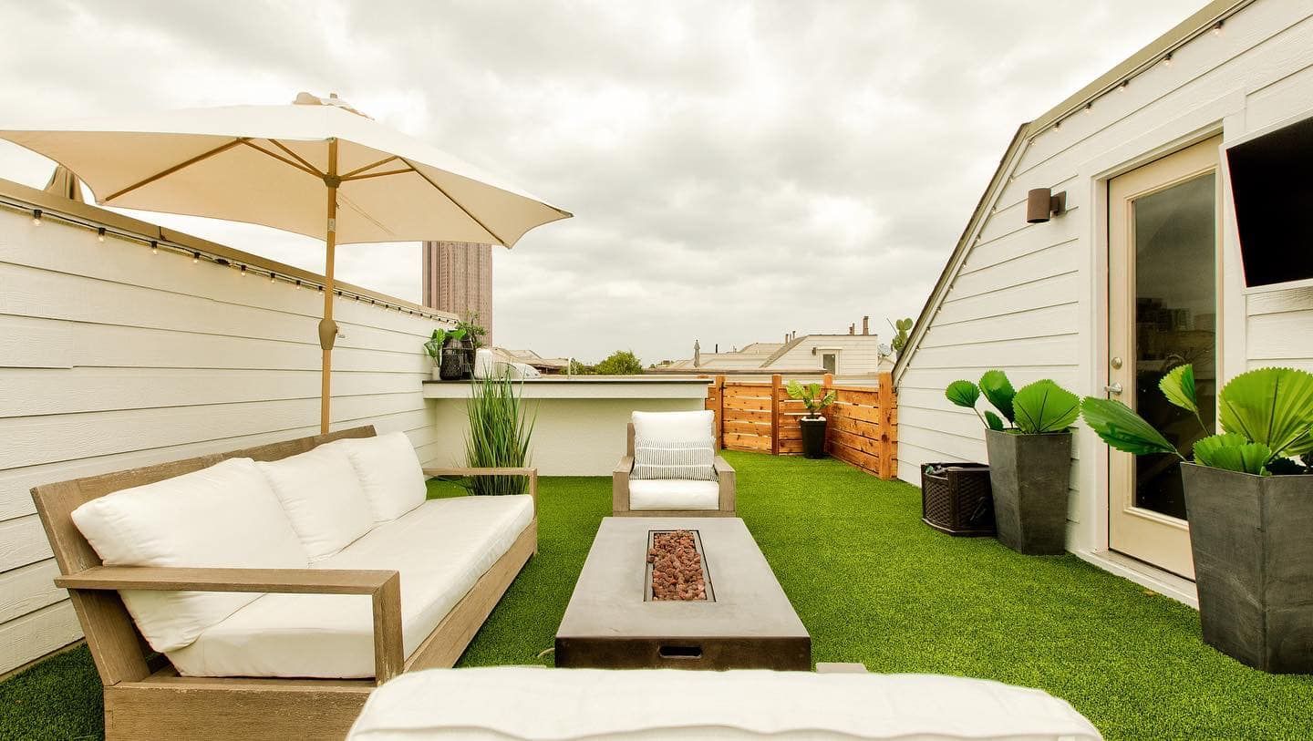 Rooftop patio with white furniture, fire pit, umbrella, artificial grass, and door. Cloudy sky.