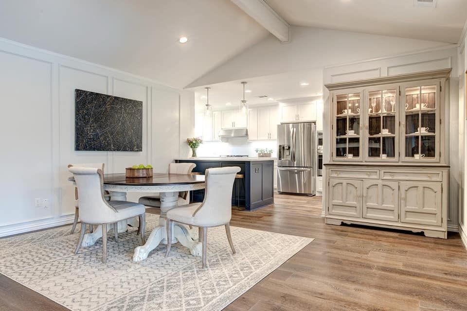 Dining room with table, chairs, rug, and hutch. Kitchen in the background. White and wood tones.