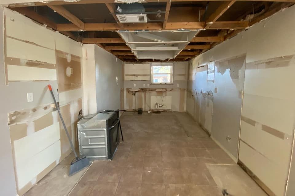 Kitchen gutted during renovation, exposed walls and ceiling, appliance visible, window in the distance.