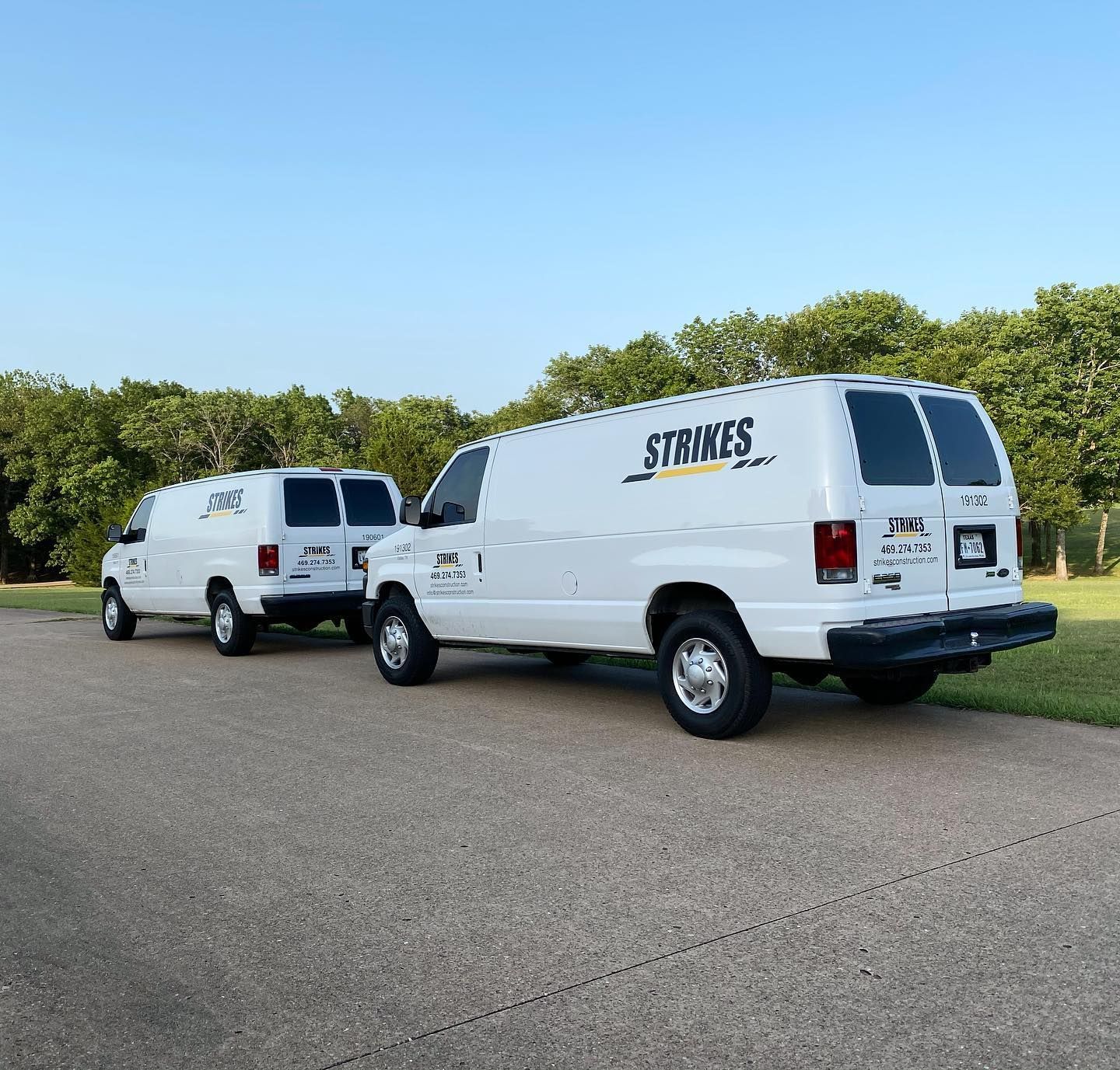 Two white vans with business logos parked on a paved road. Trees and blue sky are in the background.