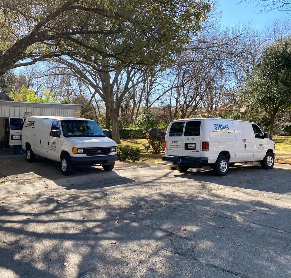 Two white vans parked in a driveway. One van is facing forward, the other backward. Trees and a house are in the background.