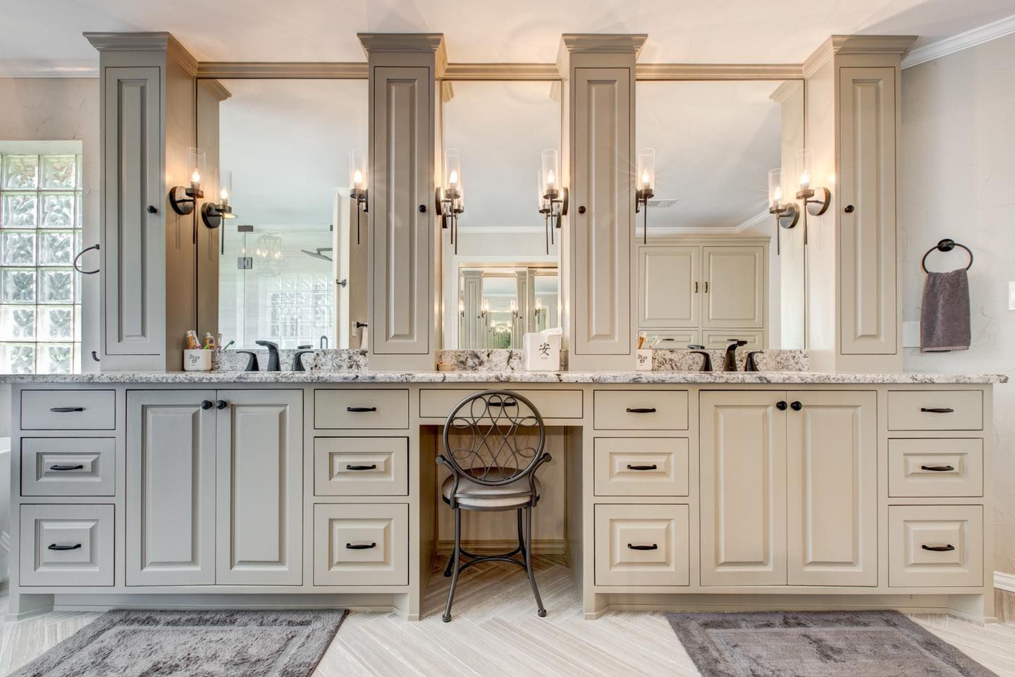 Bathroom with triple vanity. Grey and cream cabinets, mirrors, and sconces. Features a central vanity chair.