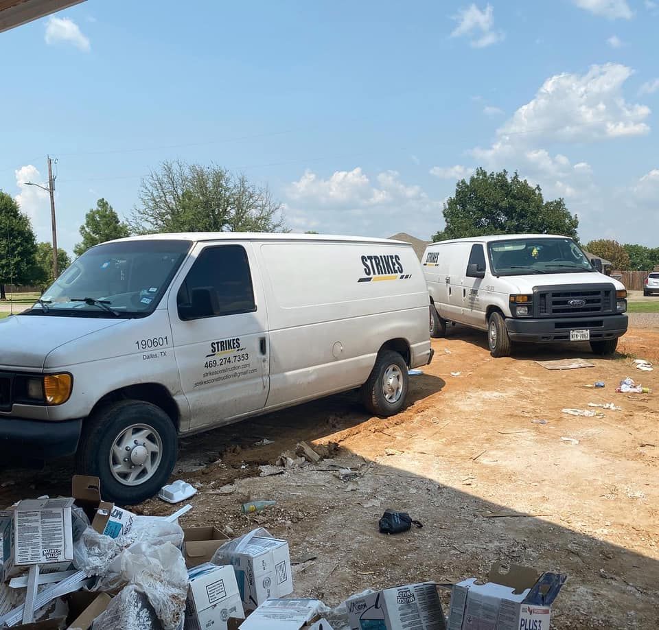 Two white work vans parked on a construction site; blue sky and trees in the background.