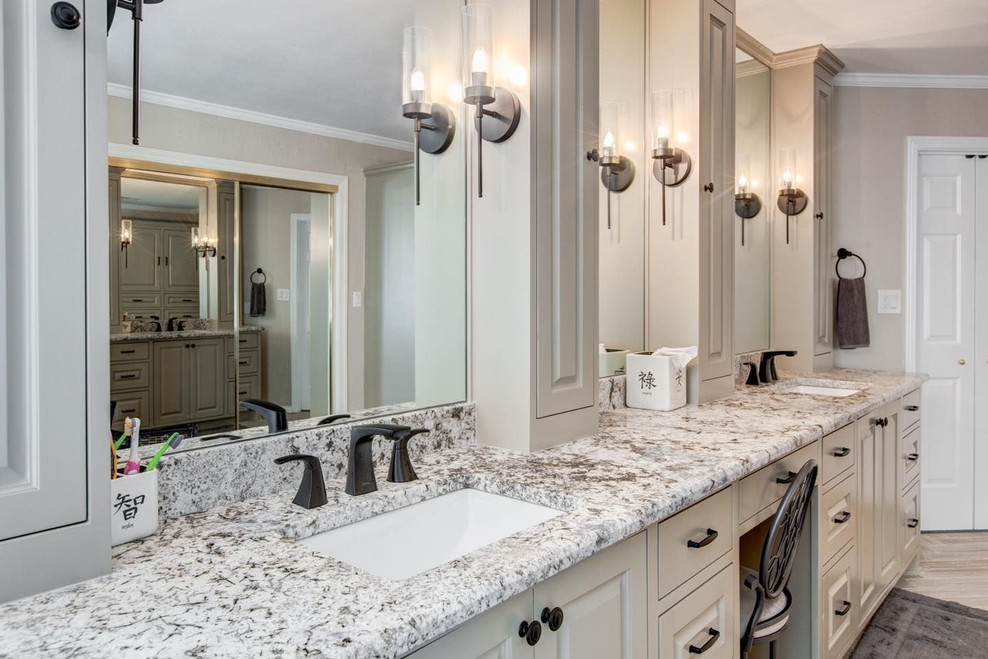 Bathroom with light gray cabinets, speckled countertop, multiple sinks, and sconce lighting.